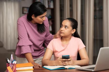 a concerned mother leans over a teenager, who is holding a phone on top of an open textbook and in front of an open laptop